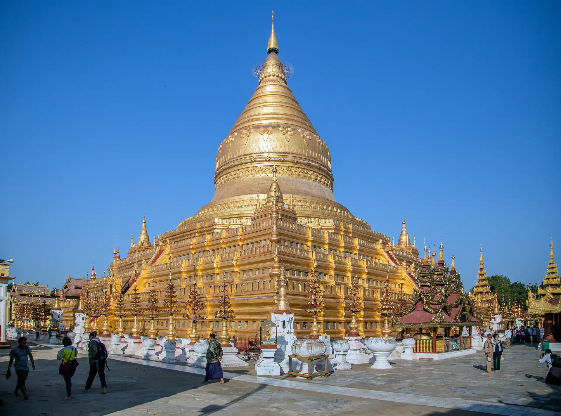 La masiva estupa dorada de la Pagoda Shwezigon brillando bajo la luz del sol, rodeada de santuarios más pequeños y devotos en vestimenta tradicional rindiendo respeto