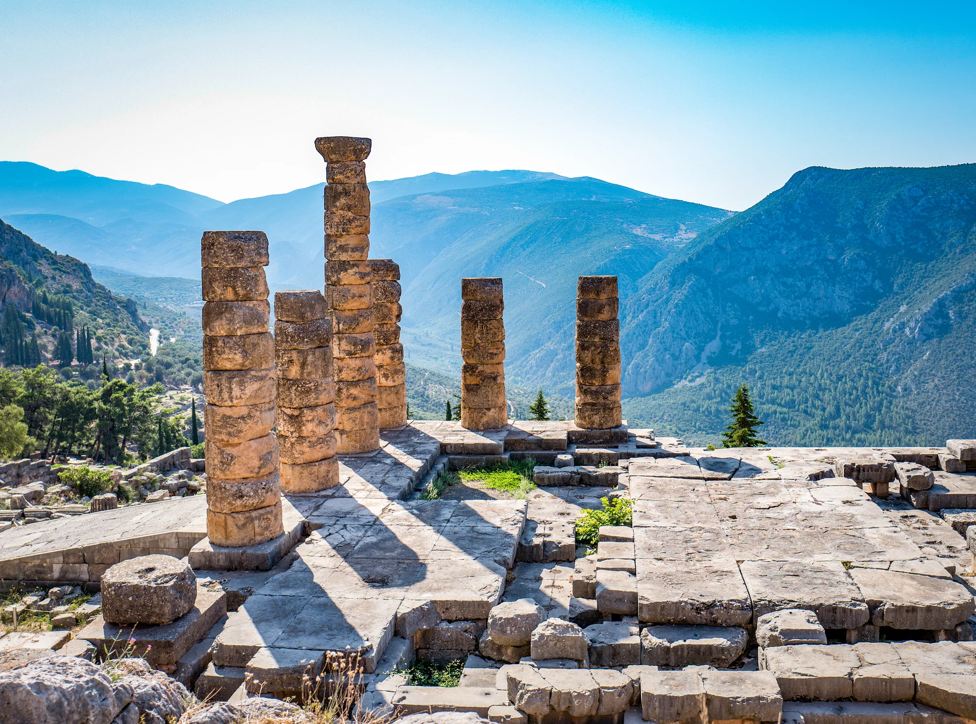 Ruins of the ancient Temple of Apollo at Delphi, showing stone foundations and columns against the backdrop of the valley of Phocis and mountains