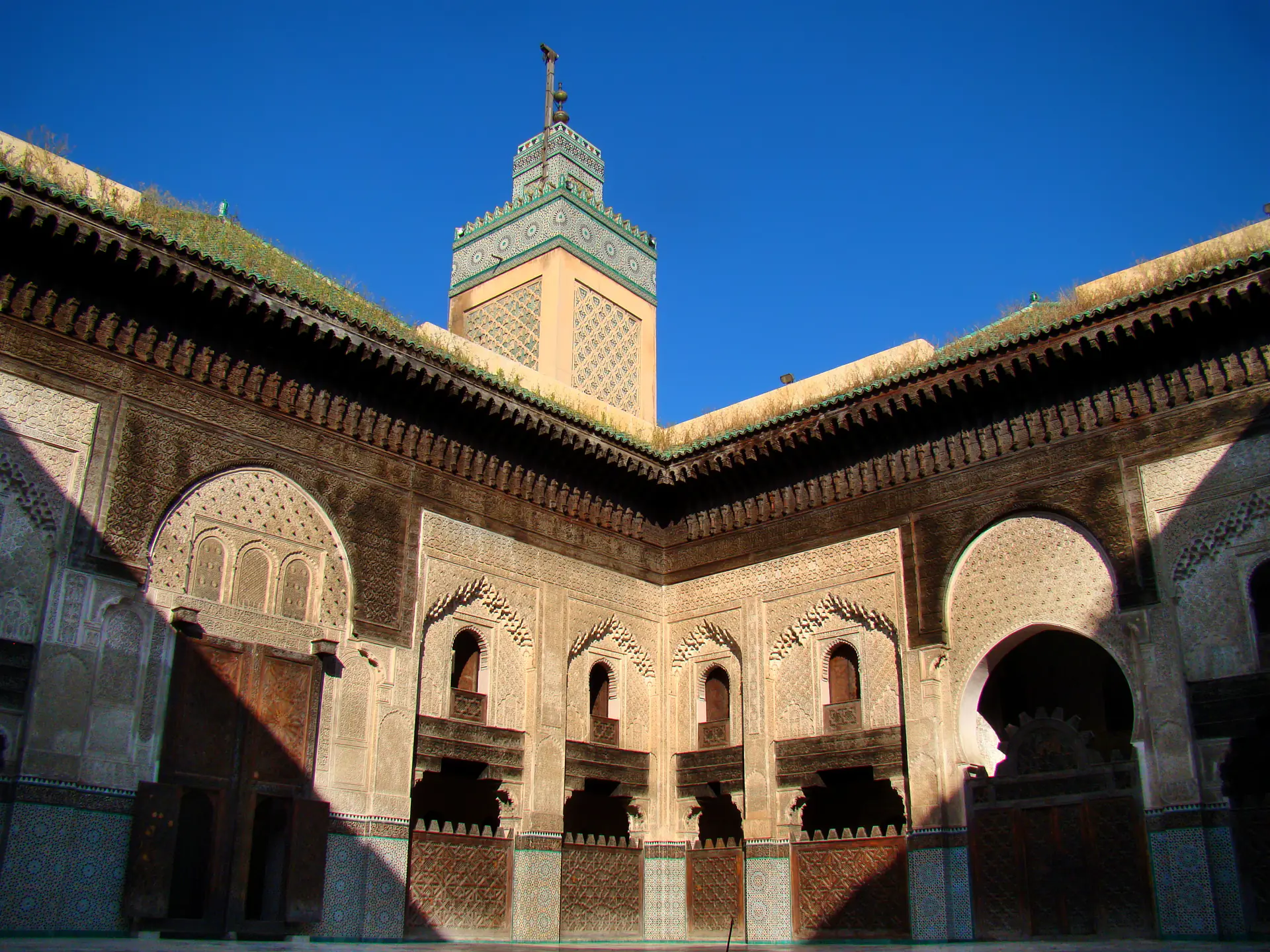 Interior courtyard of Bou Inania Madrasa displaying intricate Islamic geometric patterns, zellige tilework, carved stucco, and ornate cedar woodwork