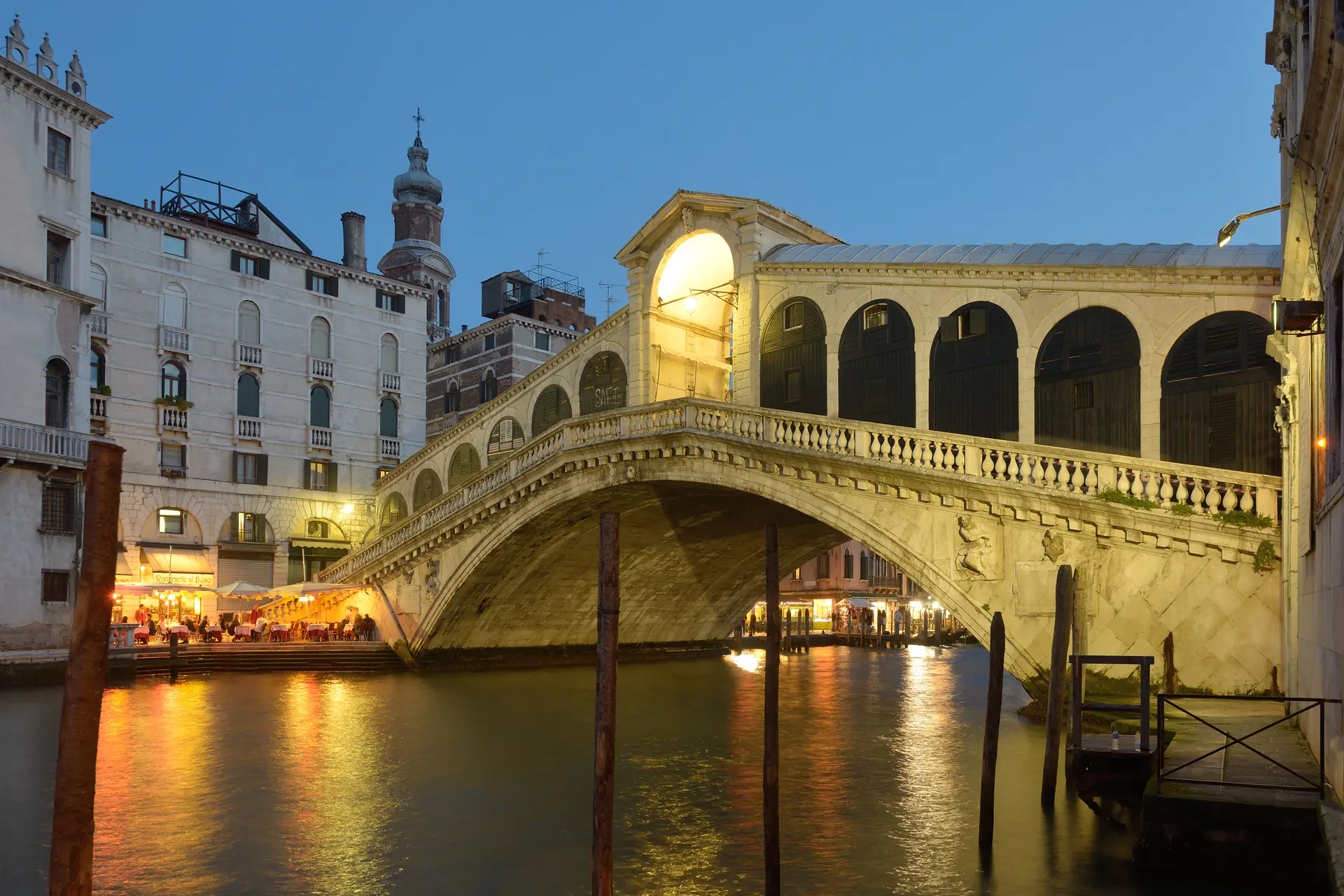 The iconic Rialto Bridge spanning the Grand Canal during blue hour, with dramatic atmospheric lighting creating deep blue tones across the water and illuminated buildings